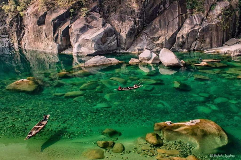 Crystal-clear waters of the Dawki river with boats gliding over visible riverbed rocks, surrounded by scenic cliffs and lush greenery